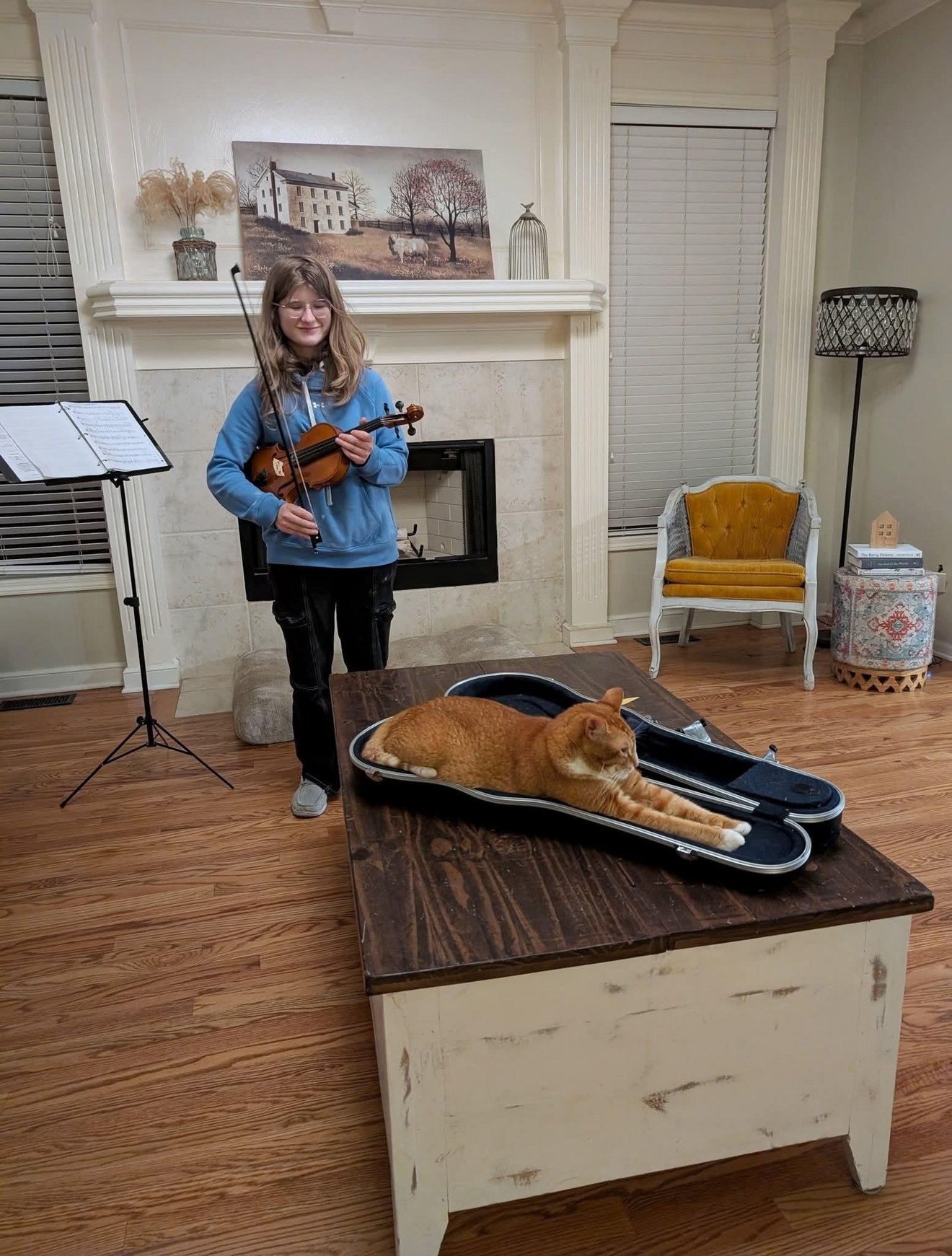 Girl playing violin while an orange tabby cat lies stretched out in an open violin case on a table
