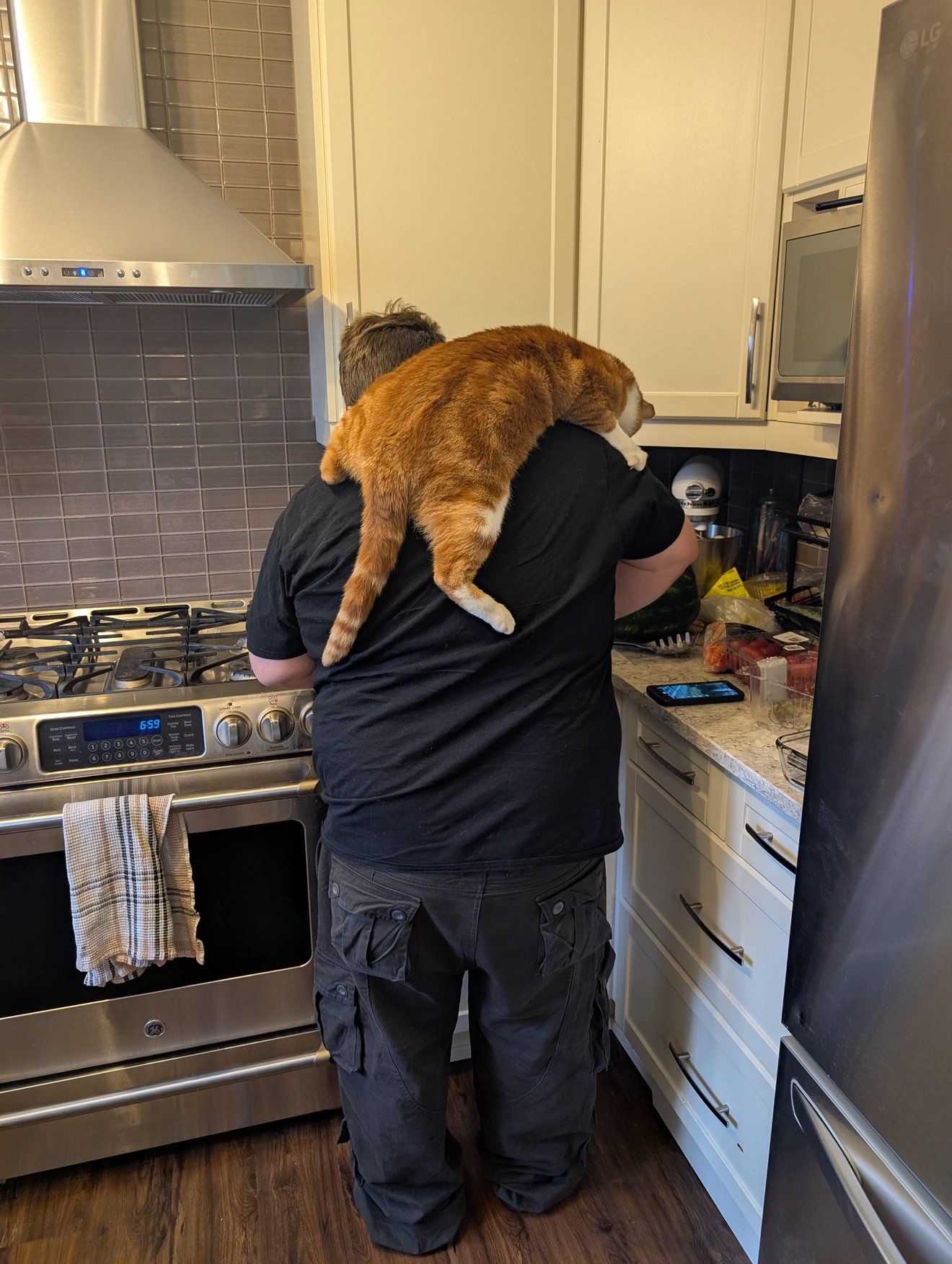 Person in black shirt holding an orange tabby cat across their shoulders while cooking in a modern kitchen
