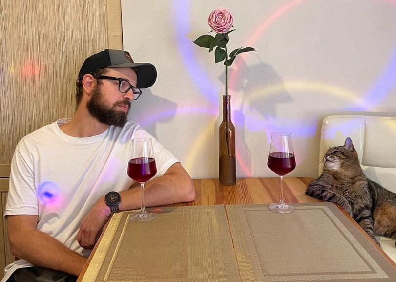 A bearded man in glasses and baseball cap sits at a wooden table with a glass of red wine and a pink rose, while a tabby cat sits beside him