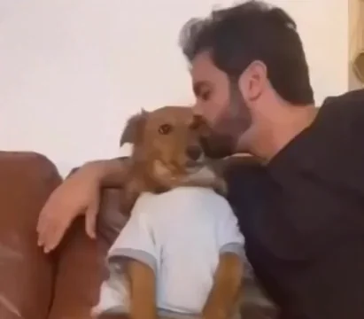 Man kissing a brown dog's face while holding it close on a couch, the dog's expression shows surprise