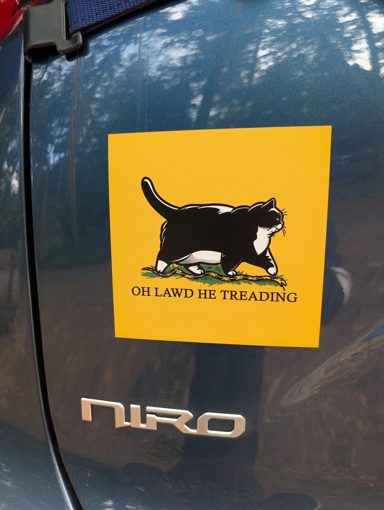 Yellow bumper sticker on dark vehicle showing black and white cat walking through grass with text reading 'oh lawd he treading'