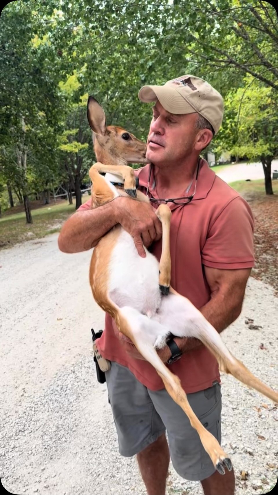 Man in baseball cap holding a tan and white deer in his arms on a gravel path lined with trees
