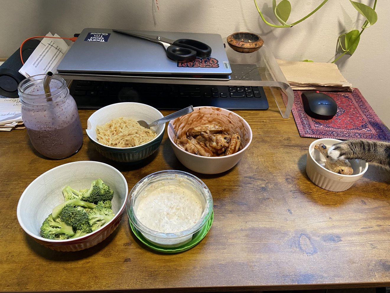 Tabby cat peeking at food bowls on desk next to laptop, noodles, broccoli, and sauces