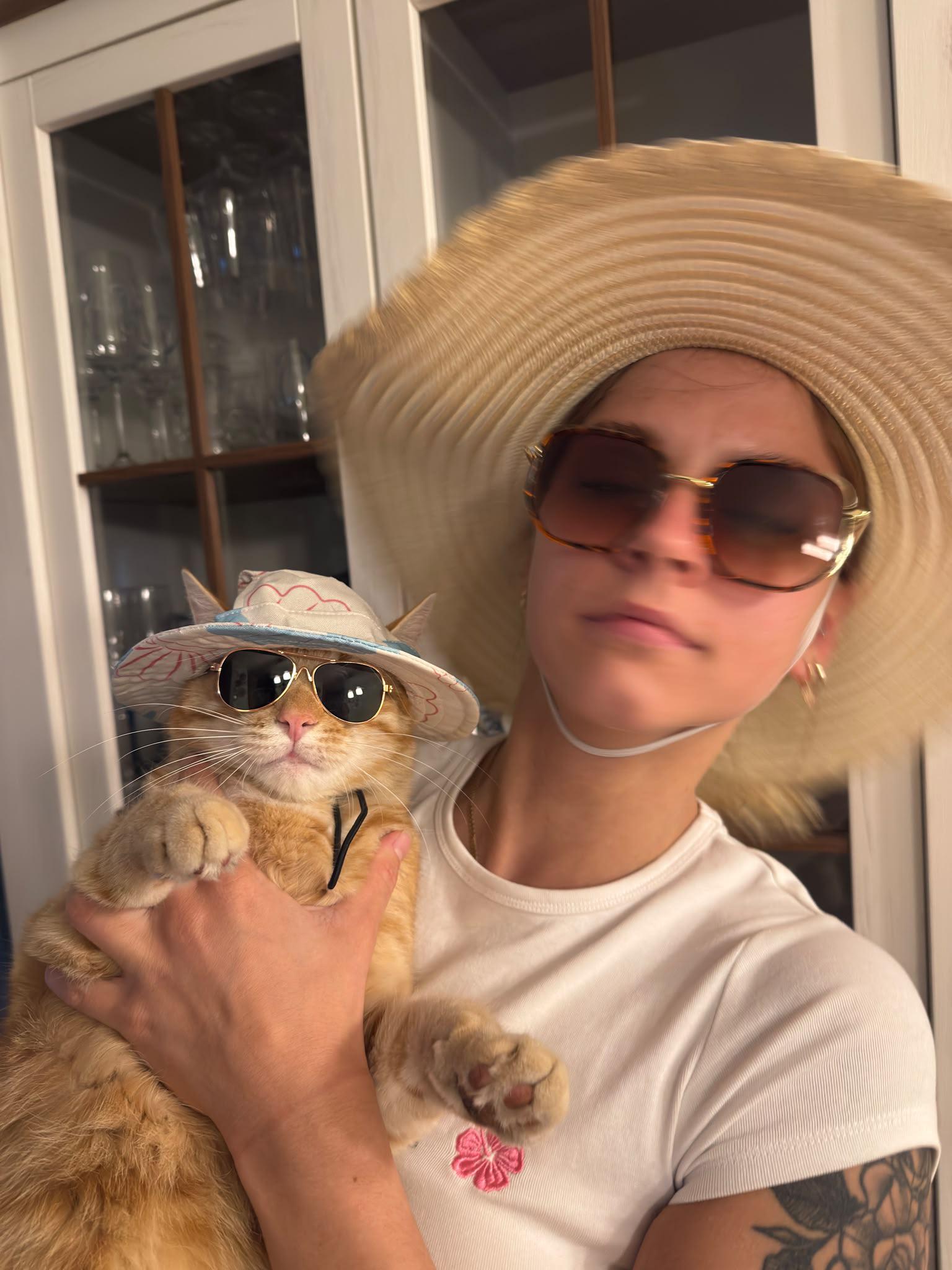 Orange tabby cat wearing sunglasses and tiny straw hat held by woman in large sun hat and sunglasses